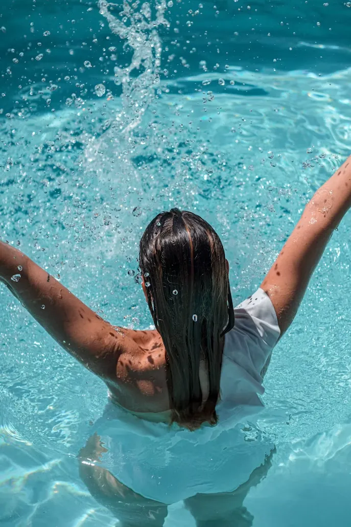 Person von hinten im türkisblauen Wasser eines Schwimmbeckens, die Arme ausgebreitet, Wassertropfen spritzen