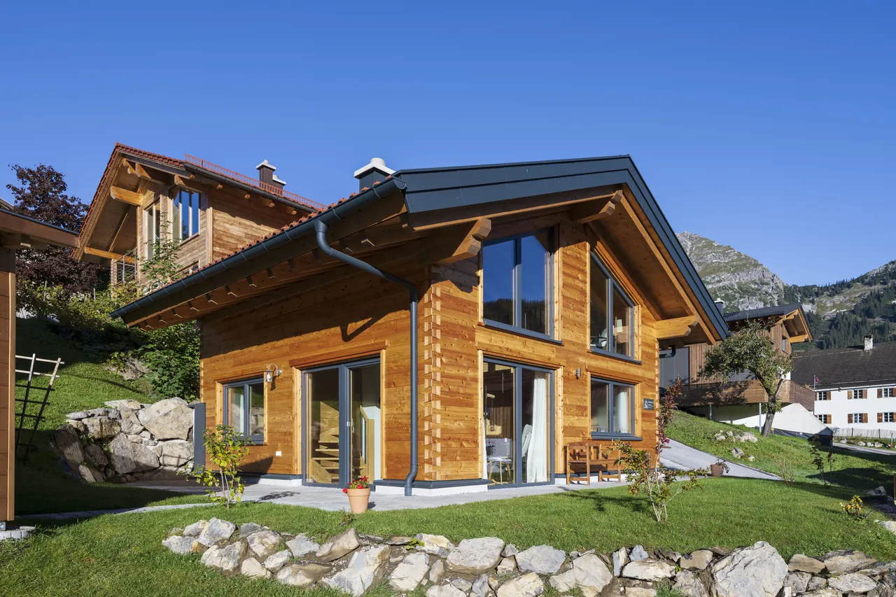 Holzhaus 171 im Sommer: Blockhaus mit großem Glasgiebel und Terrasse, dahinter Berge
