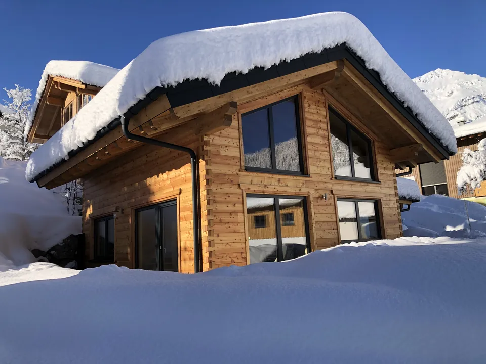 Holzhaus 171 in Berwang im Winter: Blockhaus mit Glasgiebel, das Dach dick verschneit, davor hoher Pulverschnee unter blauem Himmel